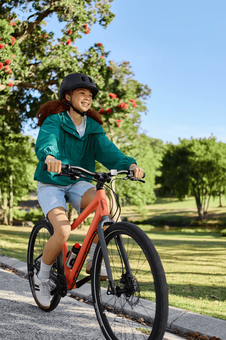 Lächelndes Mädchen mit woom READY Helm auf einem woom EXPLORE in Neon Coral, das durch einen sonnigen Park fährt.