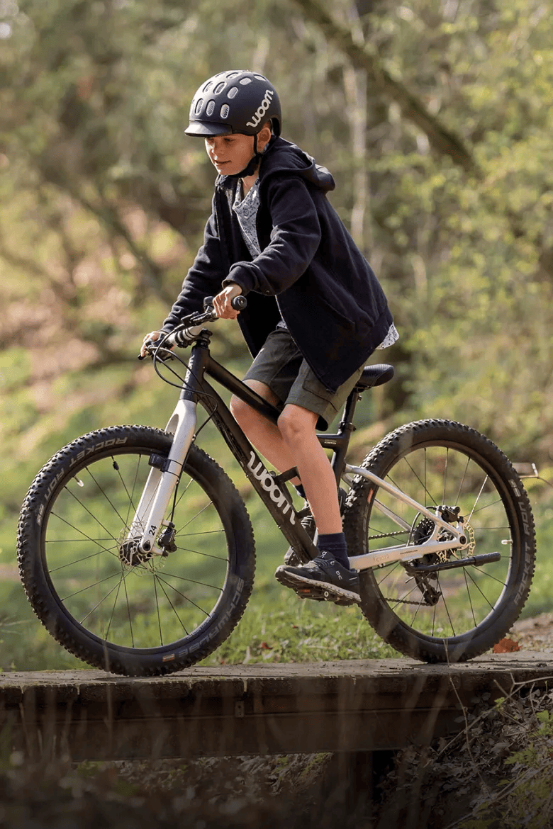 Un jeune garçon portant un casque KIDS noir roule avec son VTT woom OFF noir dans la forêt.