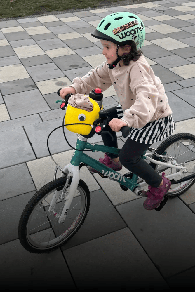 Kind mit grünem READY Helm fährt mit einem grünen woom GO bike, am Lenker ist ein gelber Fahrradkorb mit Wackelauge montiert