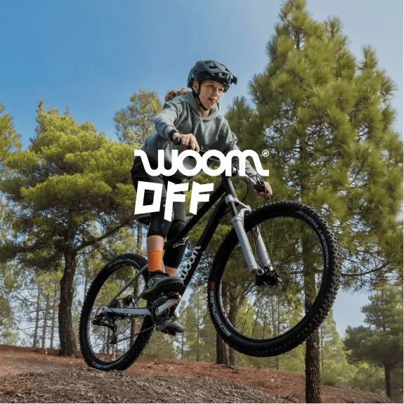 An image of a girl wearing a helmet and beginning a jump on a black woom OFF bike in a forested area.