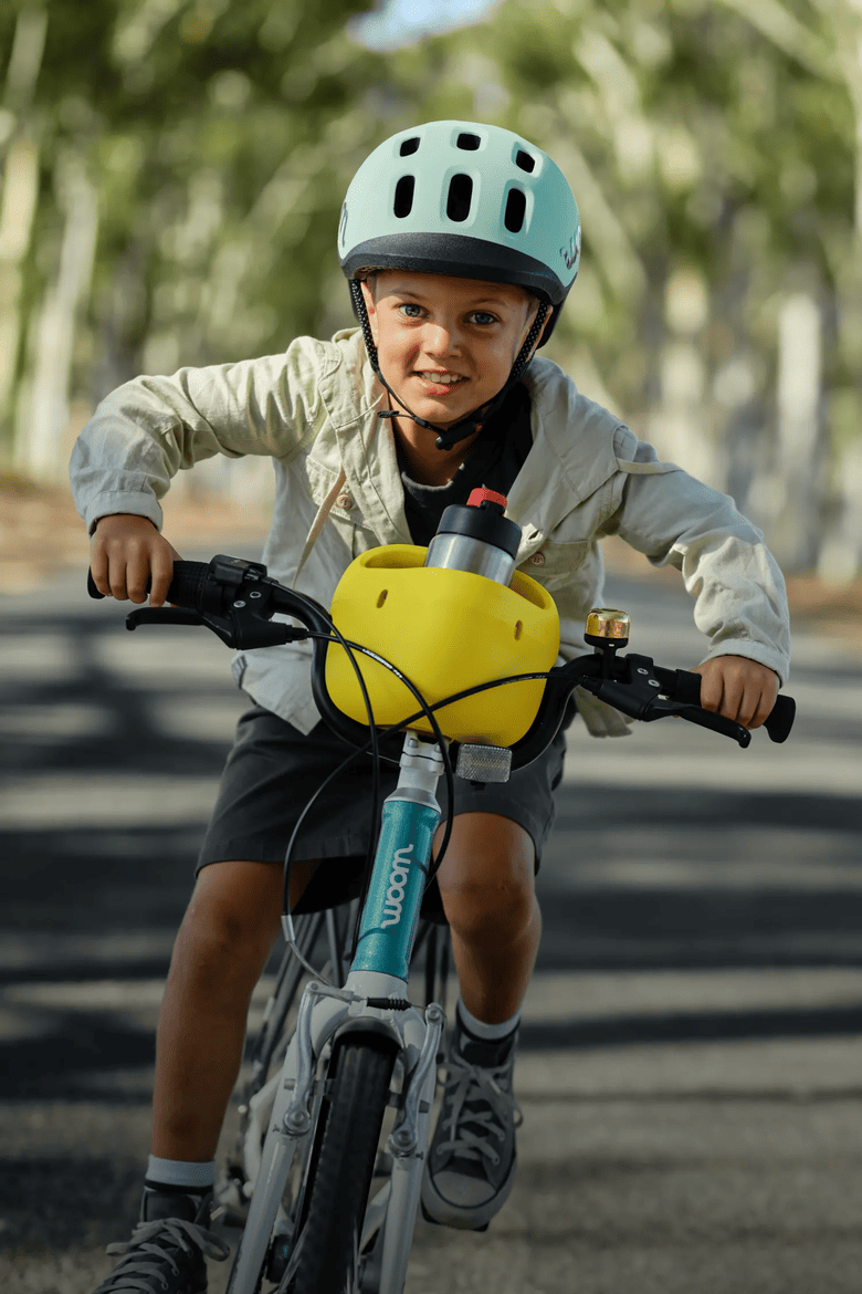 A boy wearing a woom READY helmet, riding a metallic turquoise woom GO bike with vibrant yellow POP basket holding a GLUG water bottle while riding on a road.